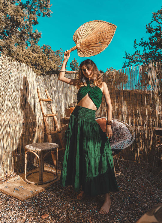 Female model is clothed in emerald halter top and tiered skirt. She holds a reed fan above her head.