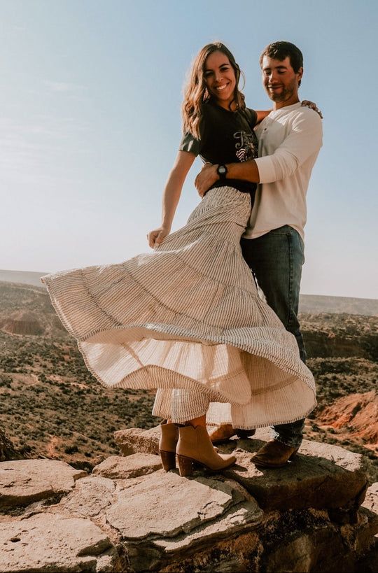 Male and female model embrace on rocks overlooking landscape. Male model is wearing white long sleeve shirt, jeans and brown boots. Female model is wearing black t-shirt, long white skirt with pinstripes and ankle boots. 