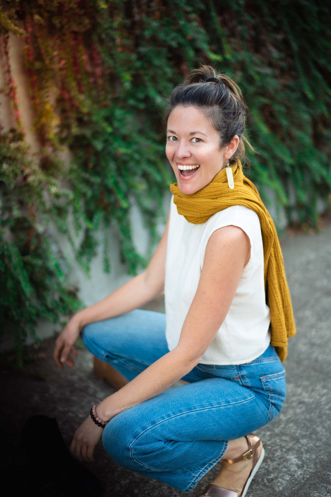 Woman crouches down on pavement wearing gold scarf draped down her back, white top and jeans.