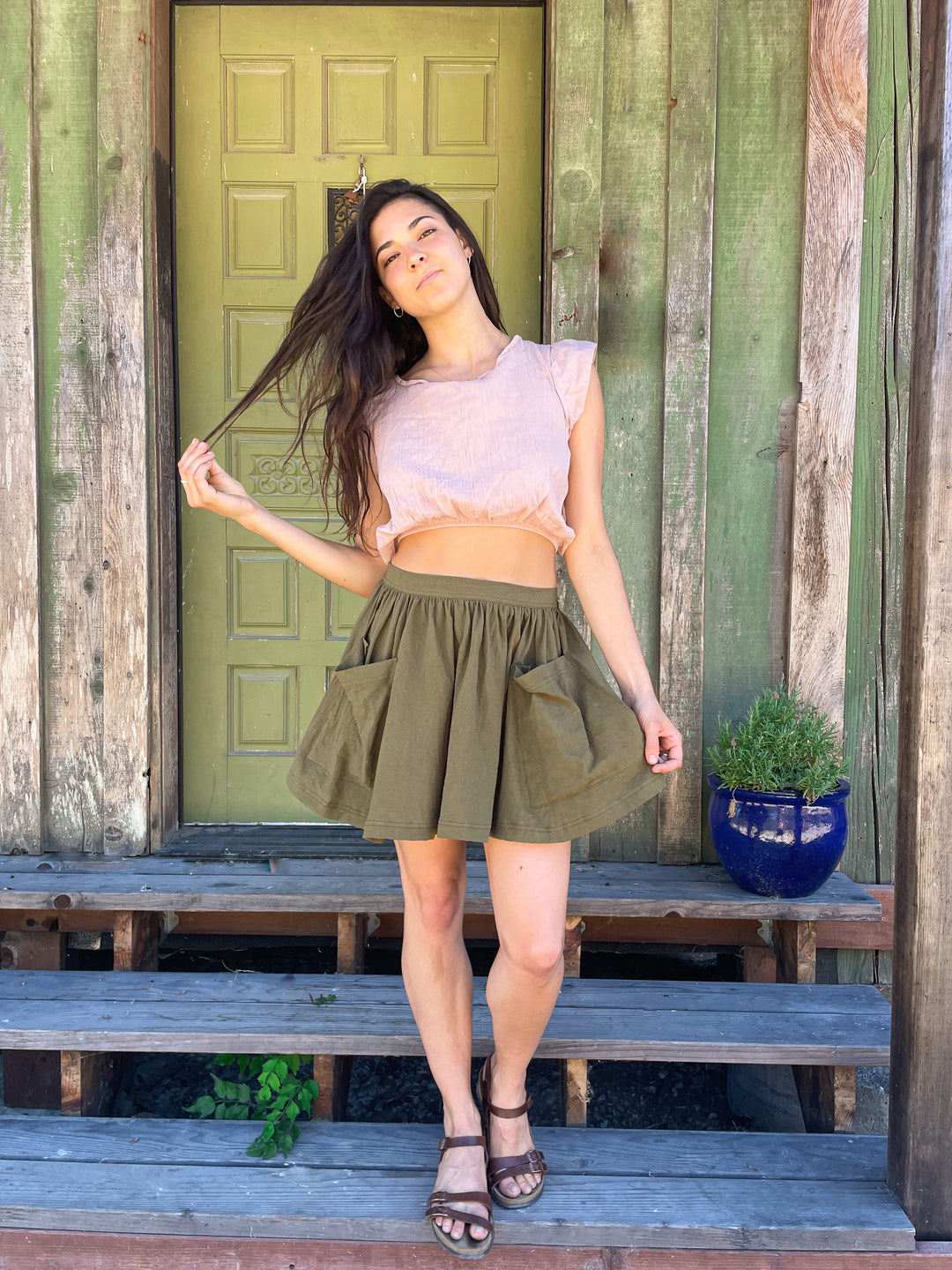 Female model poses in lightweight pink crop top paired with short dark green skirt and brown sandals.