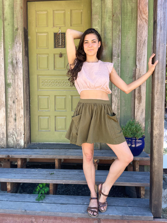 Model stands on wooden steps in front of a door wearing soft pink cotton shirt and green mini skirt with brown sandals.