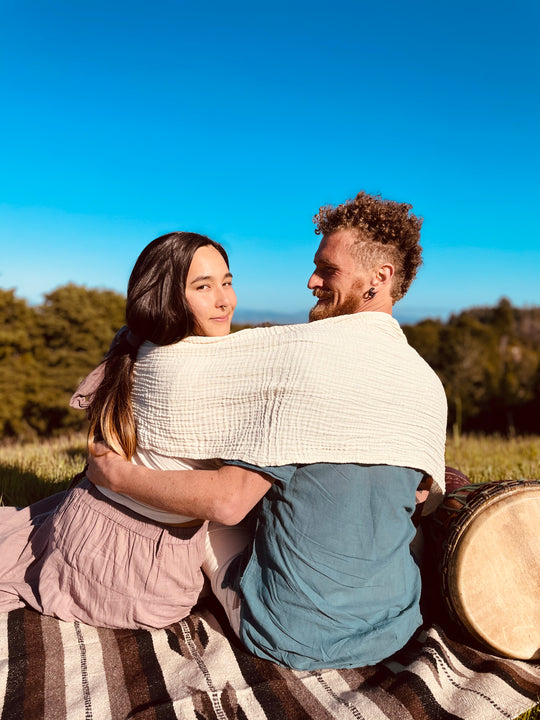 Man and woman sit outside on blanket. Creme gauze wrap is over both their shoulders. Man has arm around woman.