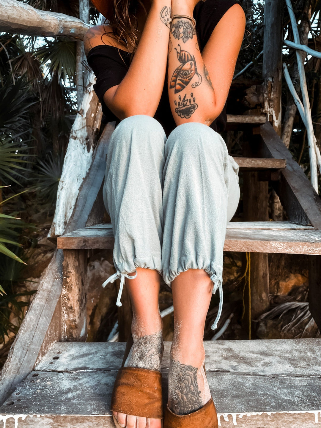 Model sits on stairs showing off light blue joggers with cuffs that have ties. Model has tattoos on feet and forearms.