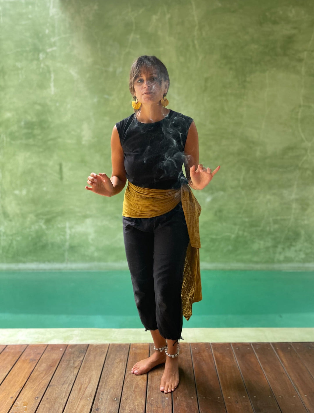 Model wears straight leg black pants, black shirt and gold fabric sash at waist. 