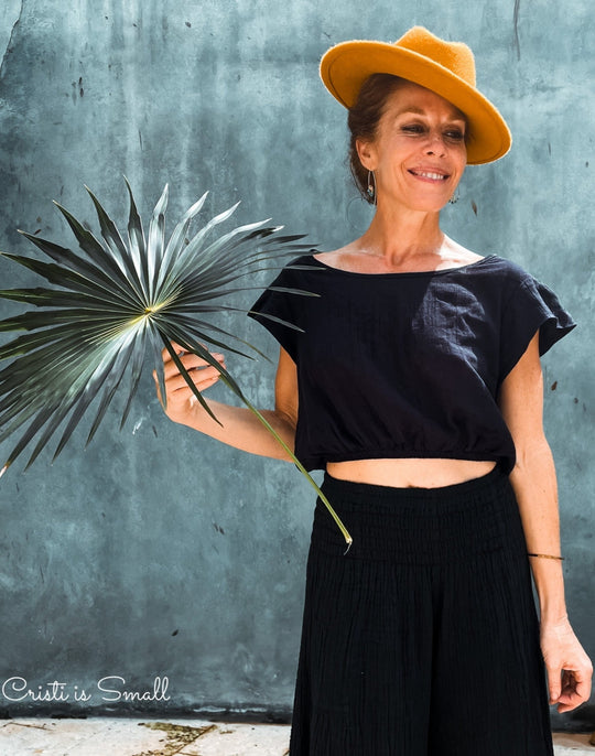 Model shows off her yellow felt hat, black crop top shirt and black bottoms. She is hold a palm in her right hand.