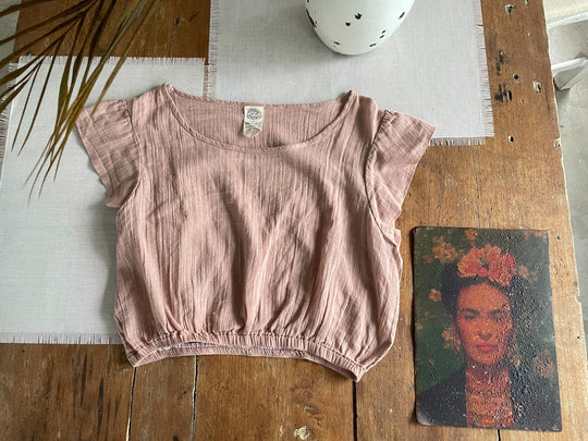 Light pink crop top shirt laid out on wood table next to picture of Frida Kahlo. 