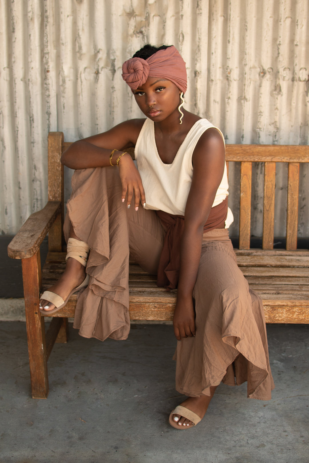Female model sits on bench with right leg up and left leg on the ground. Her right arm is resting on her right leg. She is wearing a head wrap, white tank crop top, wrap at waist, bell bottom pants and sandals.
