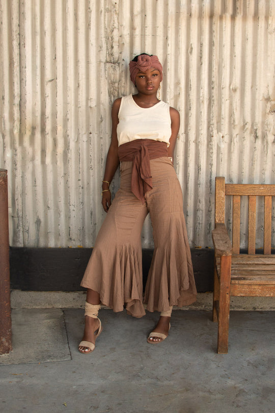 Woman leans against corrugated white wall. There is a bench to her right. She is wearing a rose-color head wrap, natural-color tank top, wrap at waist and bell-bottom pants. She is also wearing sandals. 
