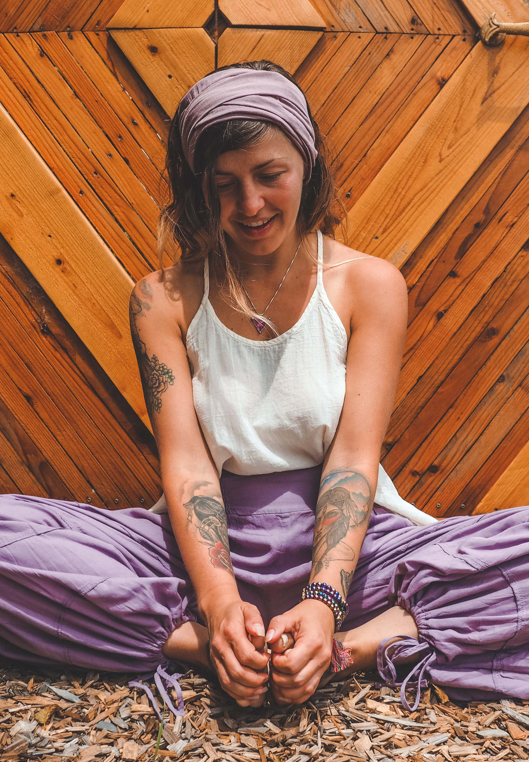 Yogini sits wearing purple head wrap, loose white tank top and purple joggers.