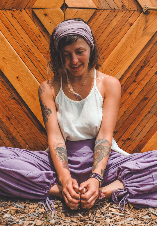 Yogini sits wearing purple head wrap, loose white tank top and purple joggers.