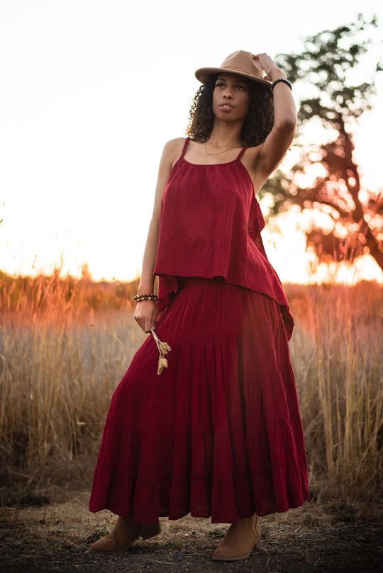 Woman is standing in field wearing brown hat, ruby red tank top, ruby red skirt and brown ankle boots.