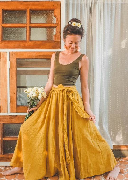 Female model stands with feet apart and looking down. She is holding daisies in her right hand. She is wearing a dark green tank crop top and long gold-color skirt with bow. Her hair is in a bun with daisies in it. 