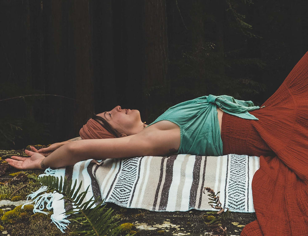 Female clothing model lays on  blanket in grass with arms above her head. She wears an orange head wrap, mint tank top tied at waist and orange bottoms. She looks peaceful.