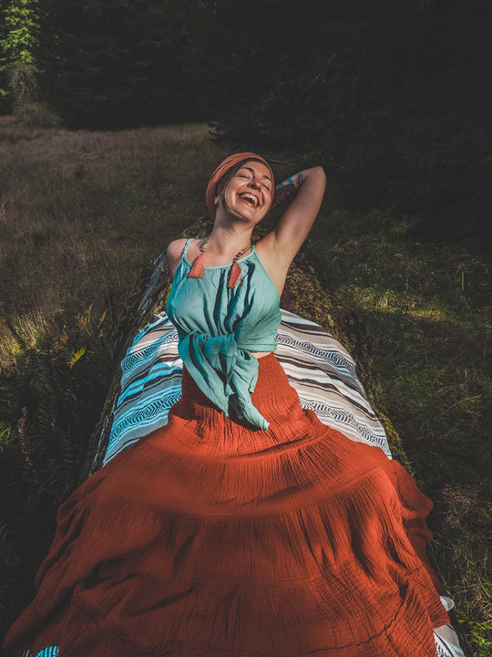 Female lays on blanket in grass with her left arm above her head. She wears an orange head wrap, aqua tank top and long orange tiered skirt.
