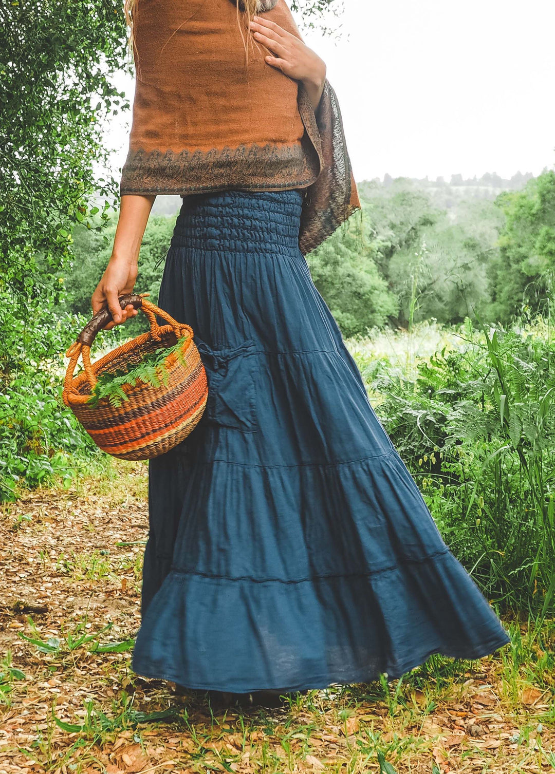 Model is wearing blue maxi skirt with side pocket and shawl. They are carrying a colorful basket in their right hand.