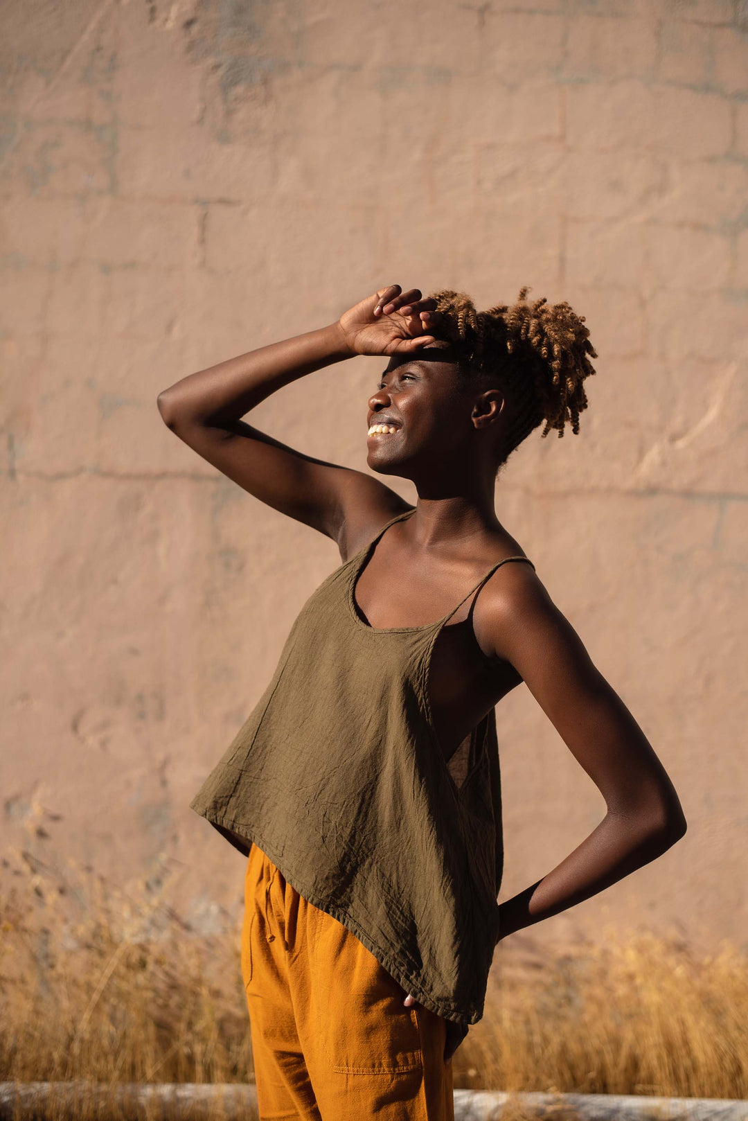 Female wearing loose green tank top and gold pants looks into the sun.