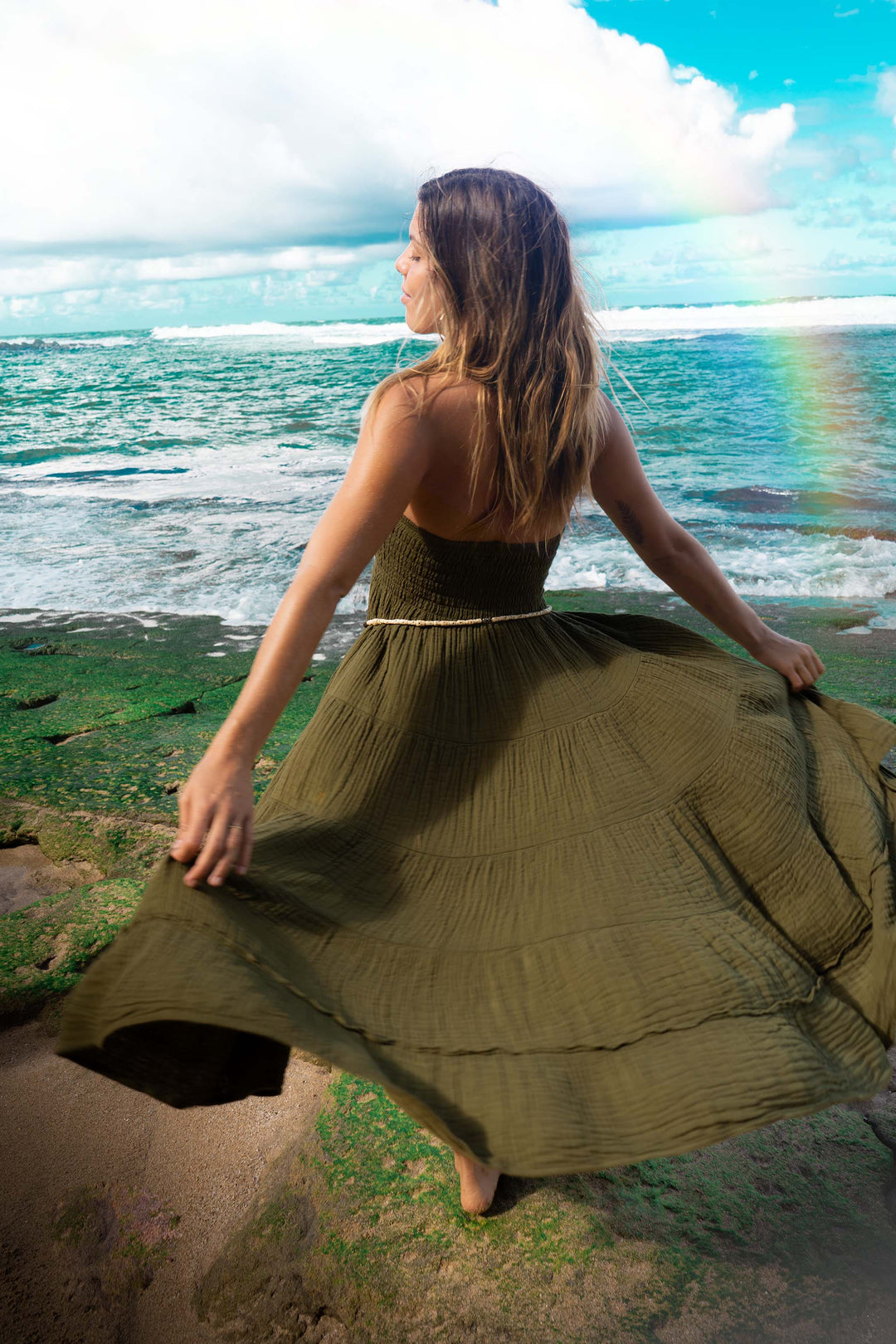 Model wears green strapless dress made of crinkle cotton. She is twirling by the ocean.