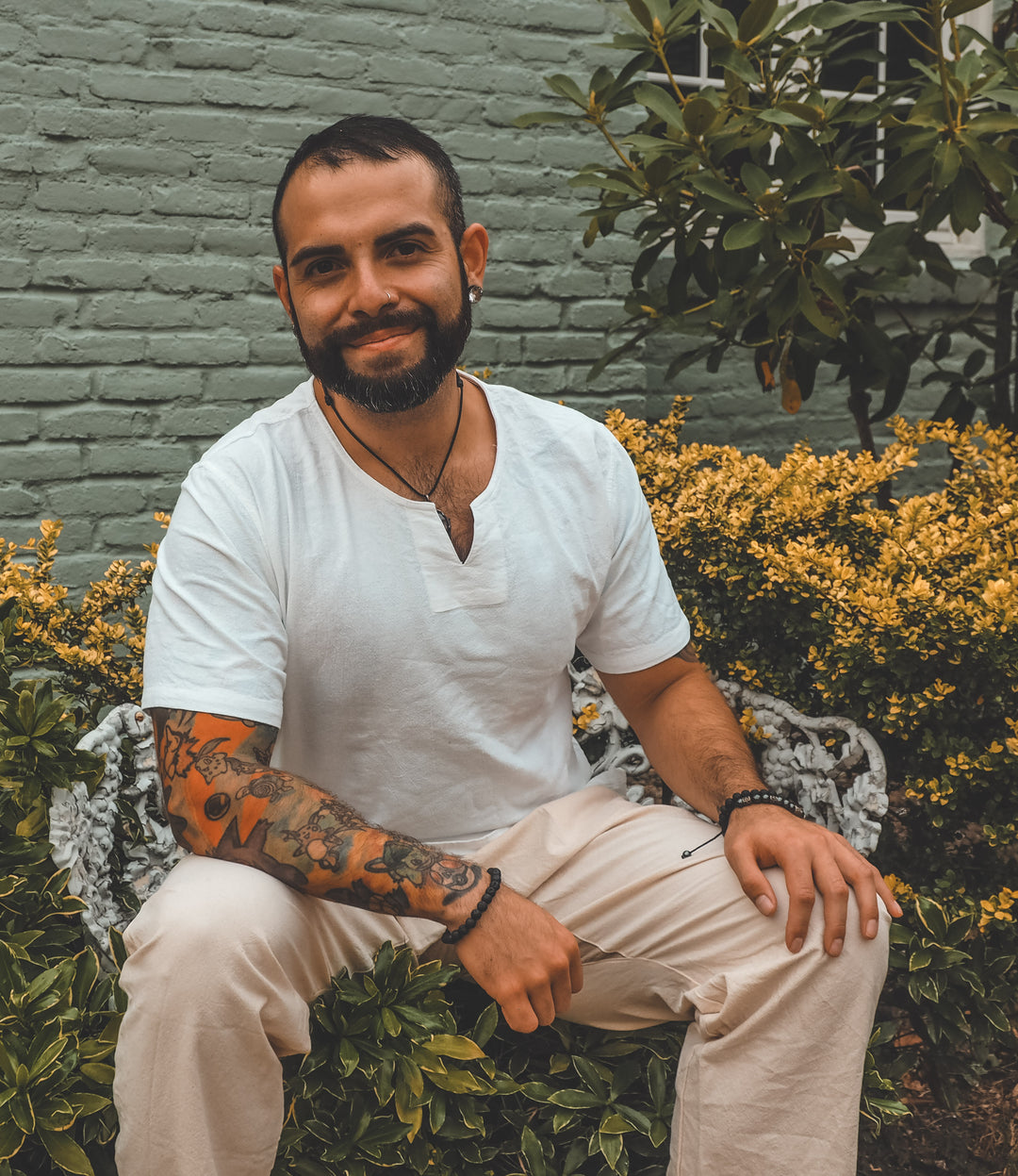 Male wears white cotton shirt with small v-neck and natural pants. He iss smiling and sitting outside in a garden.