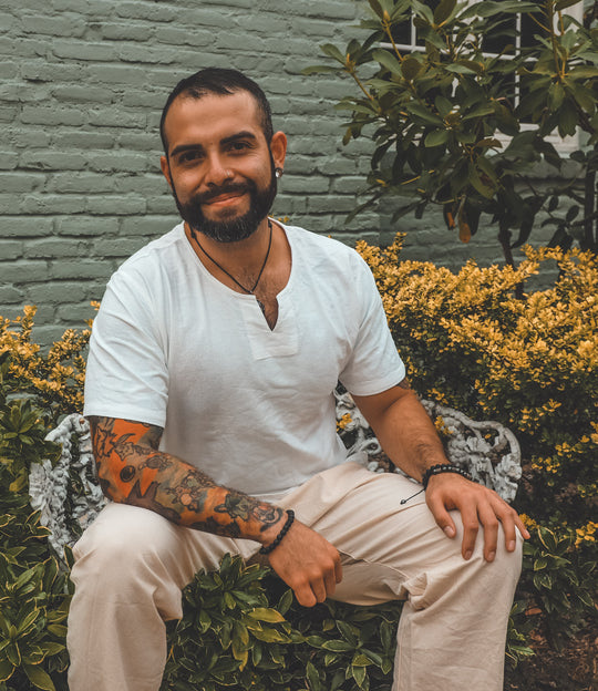Male wears white cotton shirt with small v-neck and natural pants. He iss smiling and sitting outside in a garden.