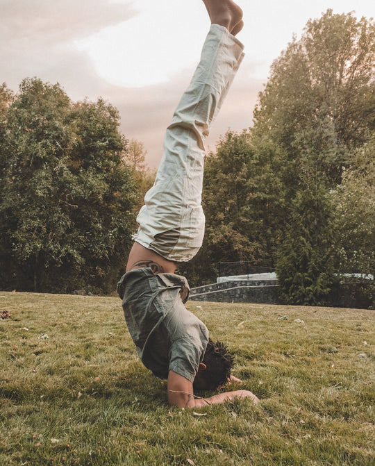 Male model is in a forearm headstand on grass. He is wearing a loose green shirt and un-dyed pants.