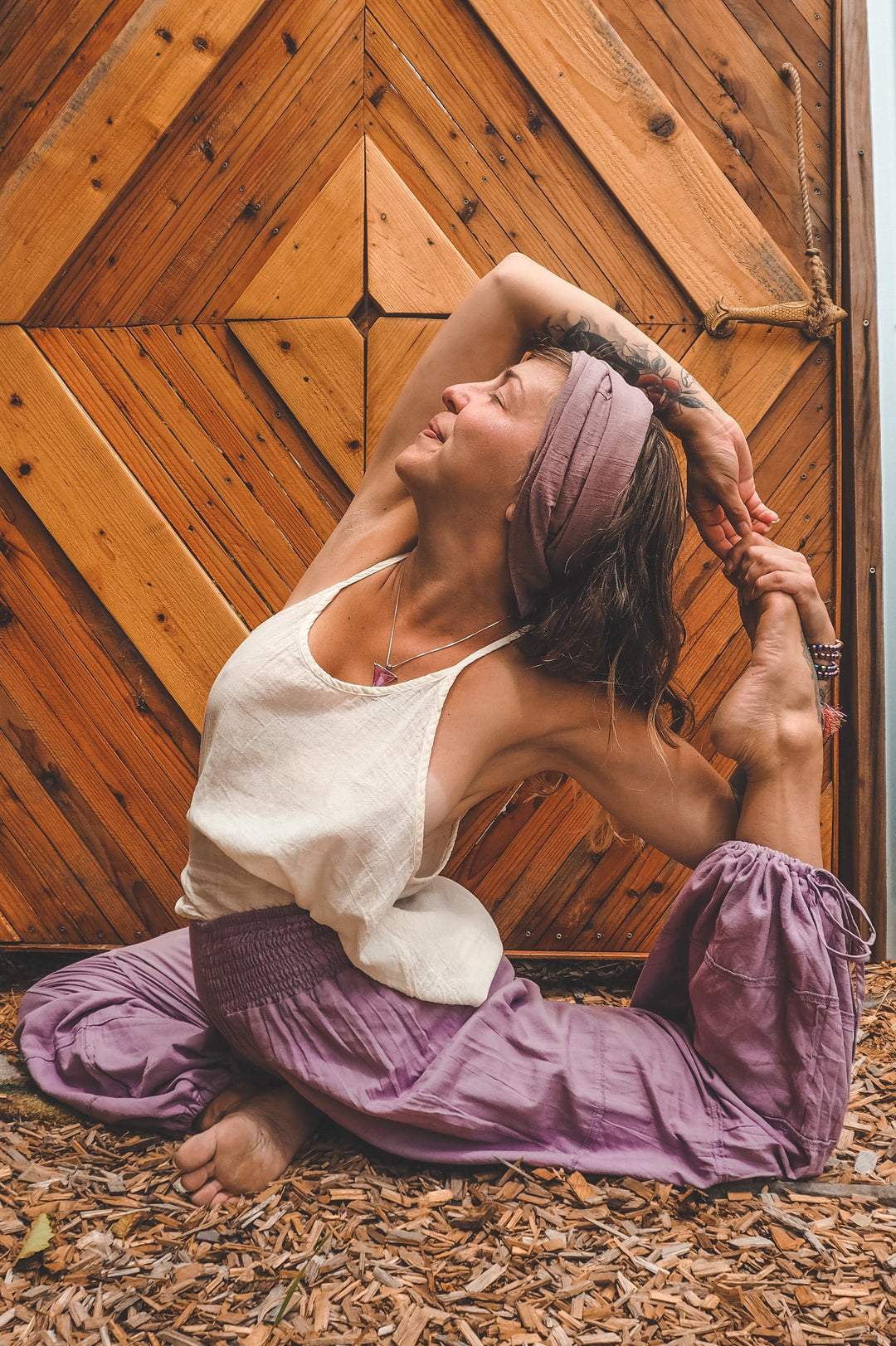 Model is in yoga pose dressed in purple head scarf, white tank top and gauze yoga pants.