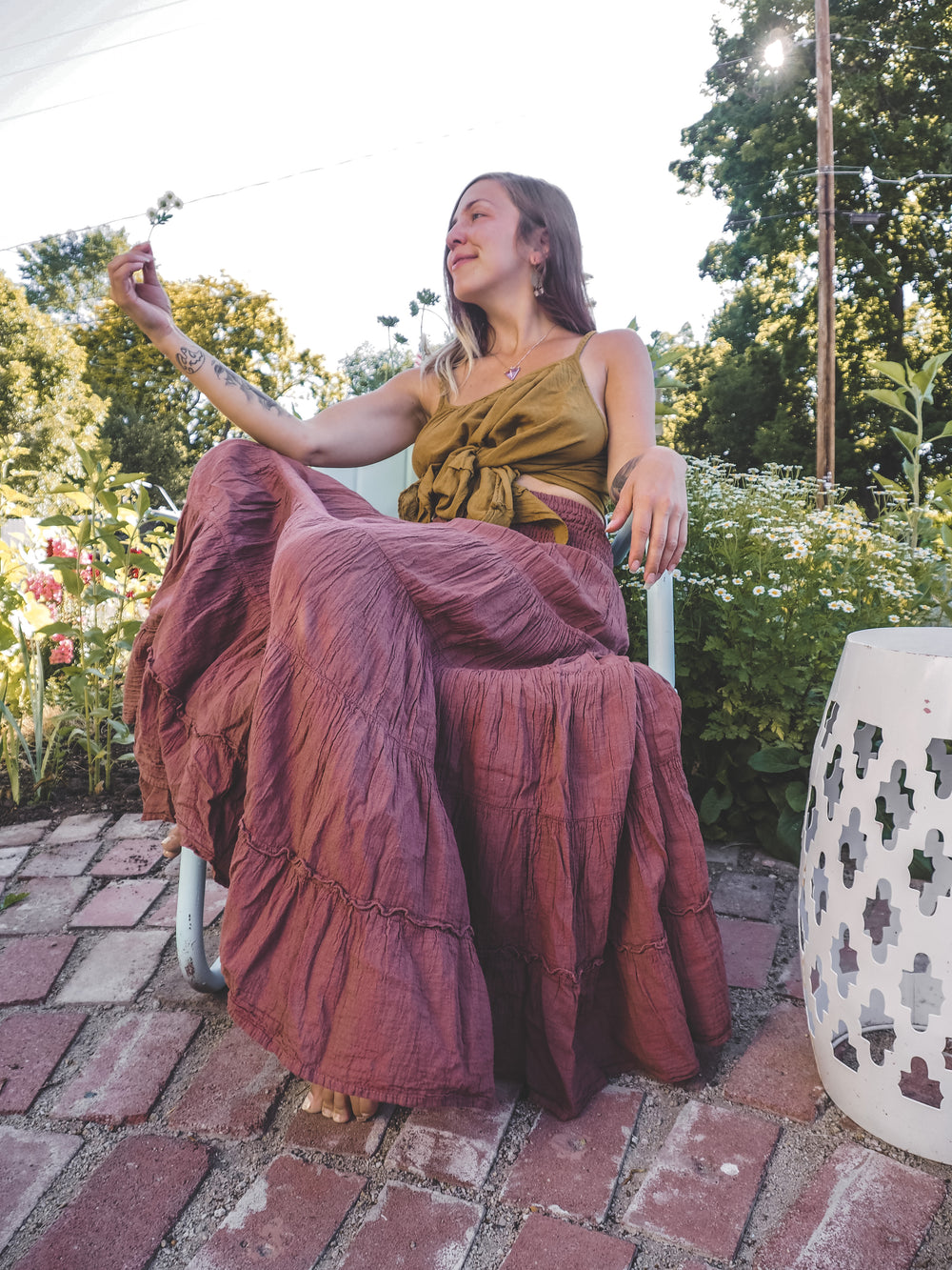 Woman sits in outside garden in a chair admiring a flower in her right hand. She is dressed in a gold-color tank top and purple maxi skirt.