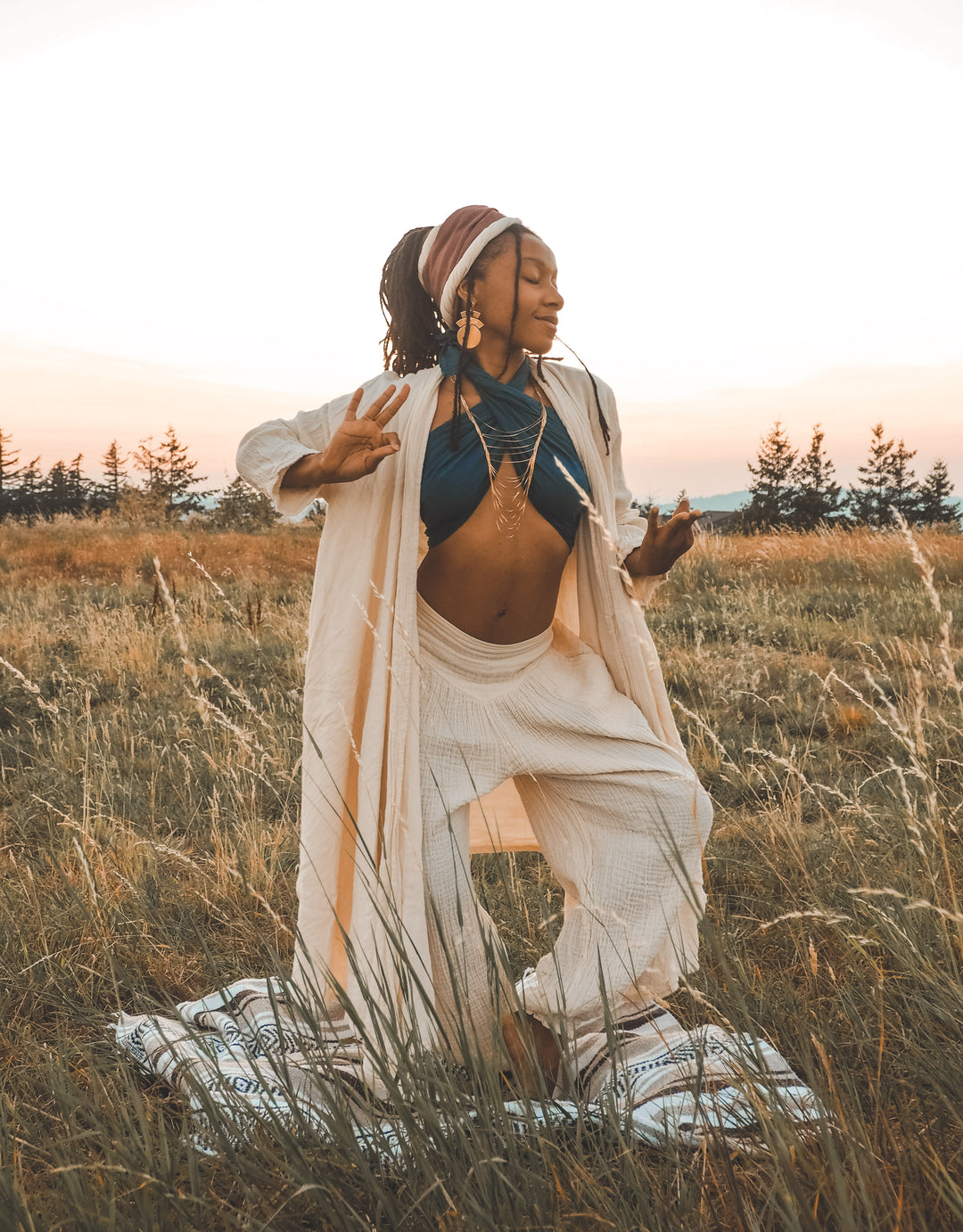 Yoga model has on wrap tied as halter top, long white cloak and loose white pants. She stands outside on a blanket.