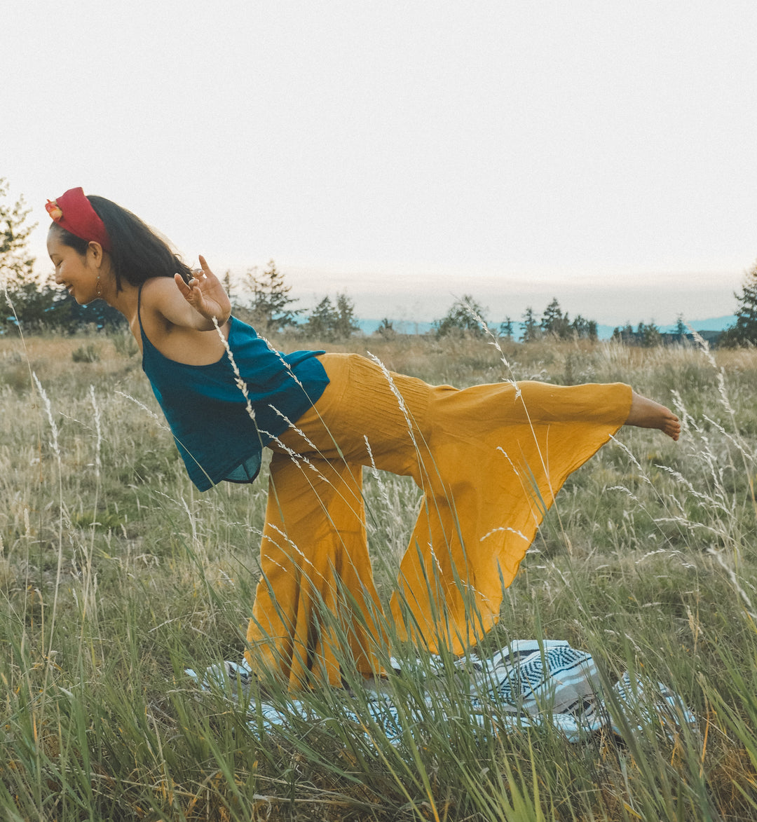 Woman is in a field wearing red head wrap, ocean blue flowy tank top and gold-color yoga pants. She balances on a blanket in warrior three yoga pose.