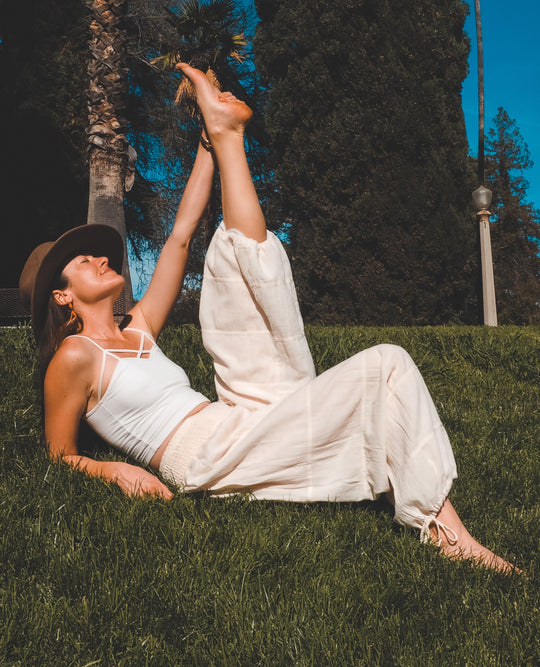 Woman lays on grass stretching left leg high. She is adorned in a white tank top and flowy white pants.