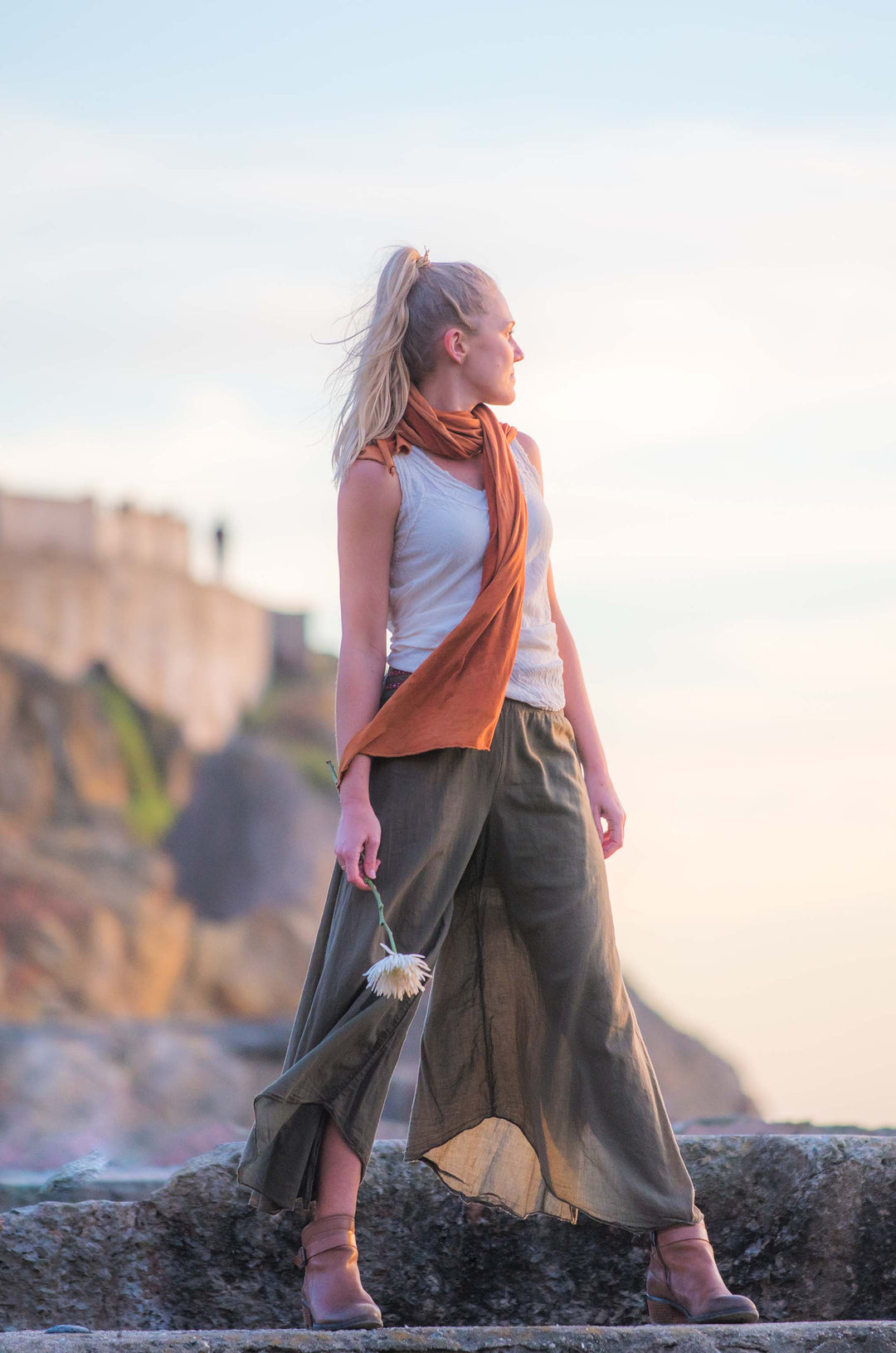 Model poses in un-dyed tank top and lightweight green pants. An orange scarf drapes around her next and down. She holds a flower in her right hand.