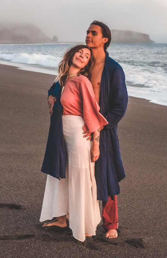 Man and woman stand on beach. Woman is wearing long pants and pink top. Man has on long cloak and pants.