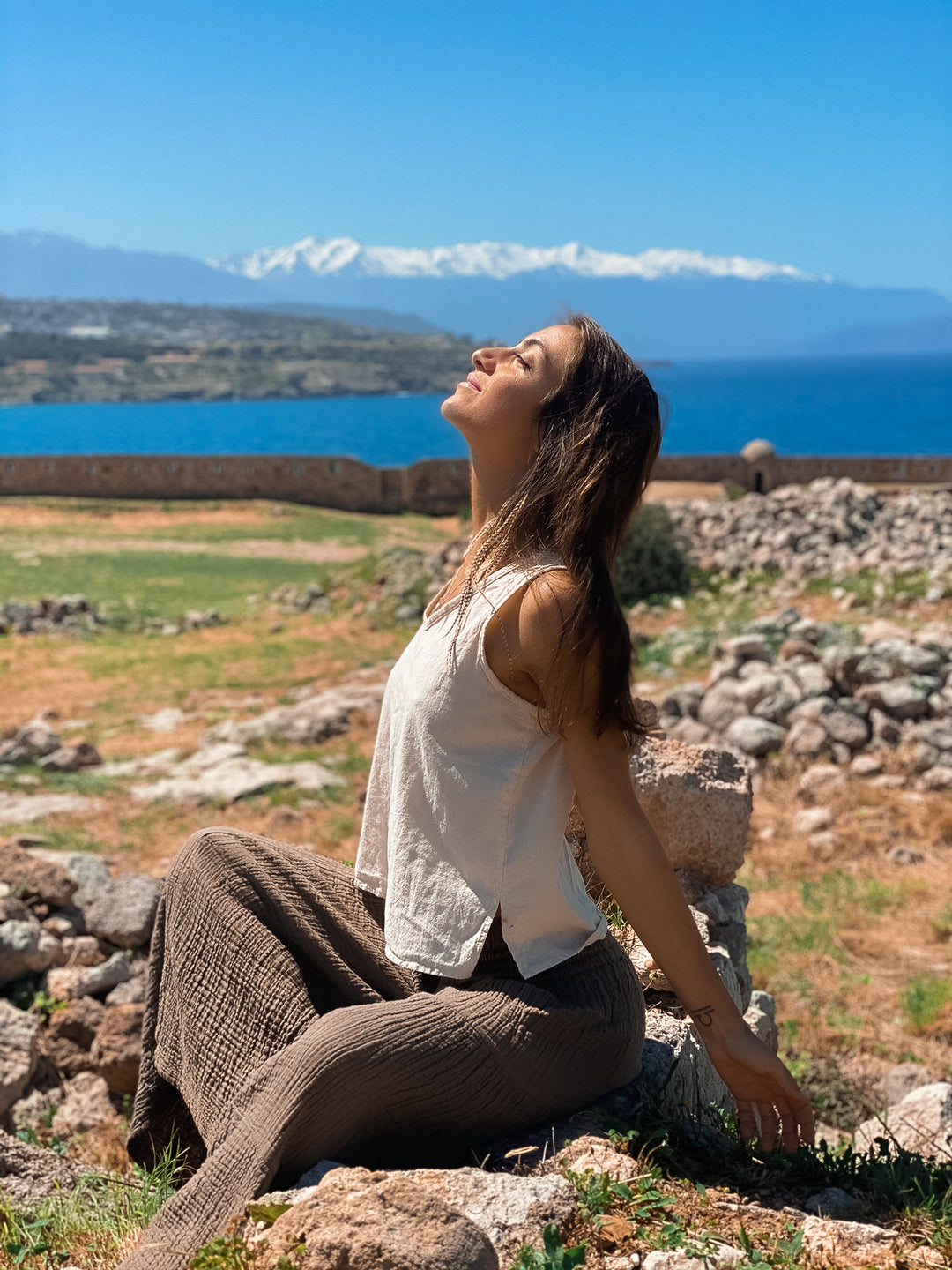 Female model sits on rocks with face angled up to the sky. There is a backdrop of green grass, rocks, water and mountains. She is wearing an un-dyed tank top and long brown gauze pants.