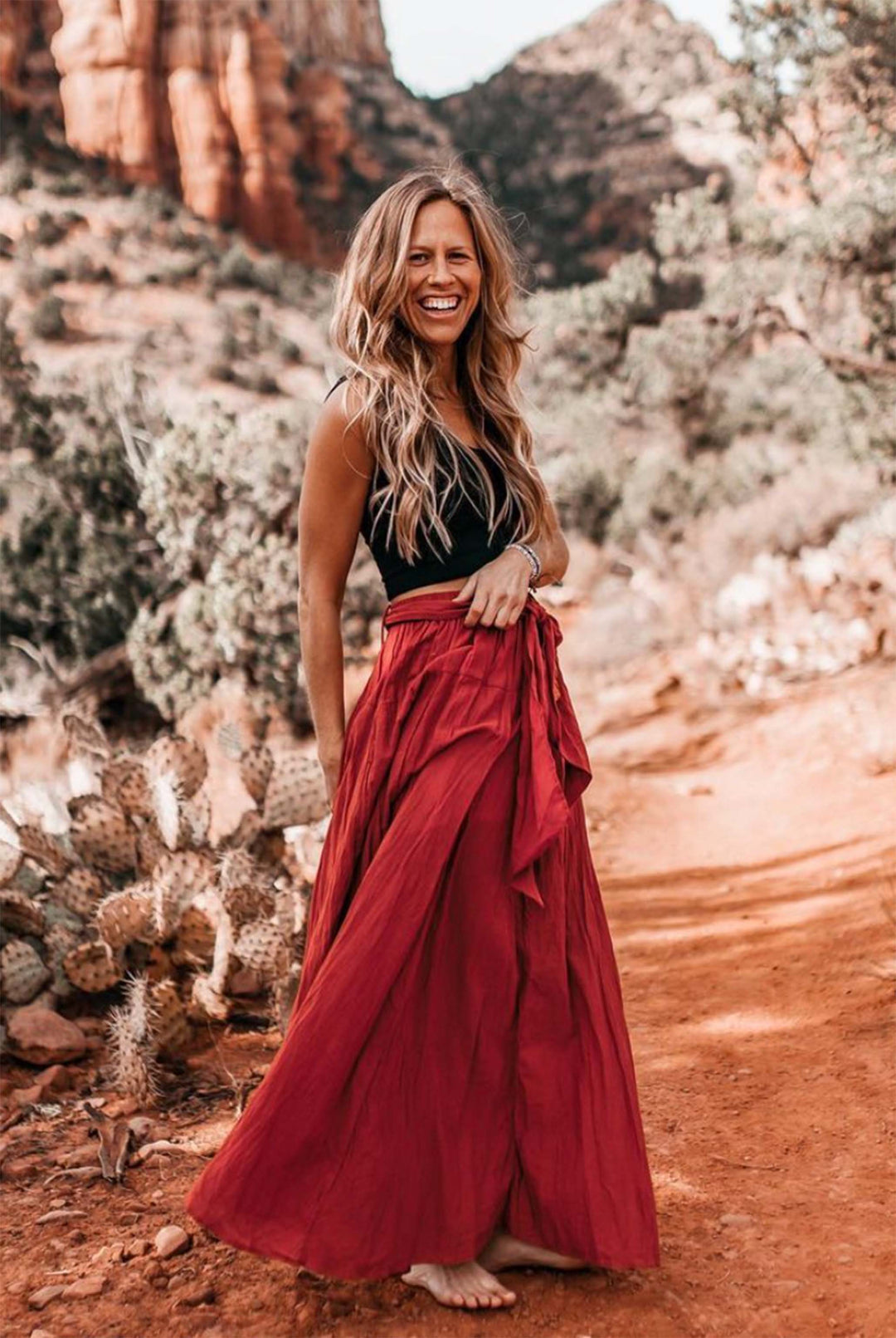 Female model standing outside with mountains and cactus in background. she is wearing a black tank crop top and long red cotton gauze skirt with bow. She is barefoot.