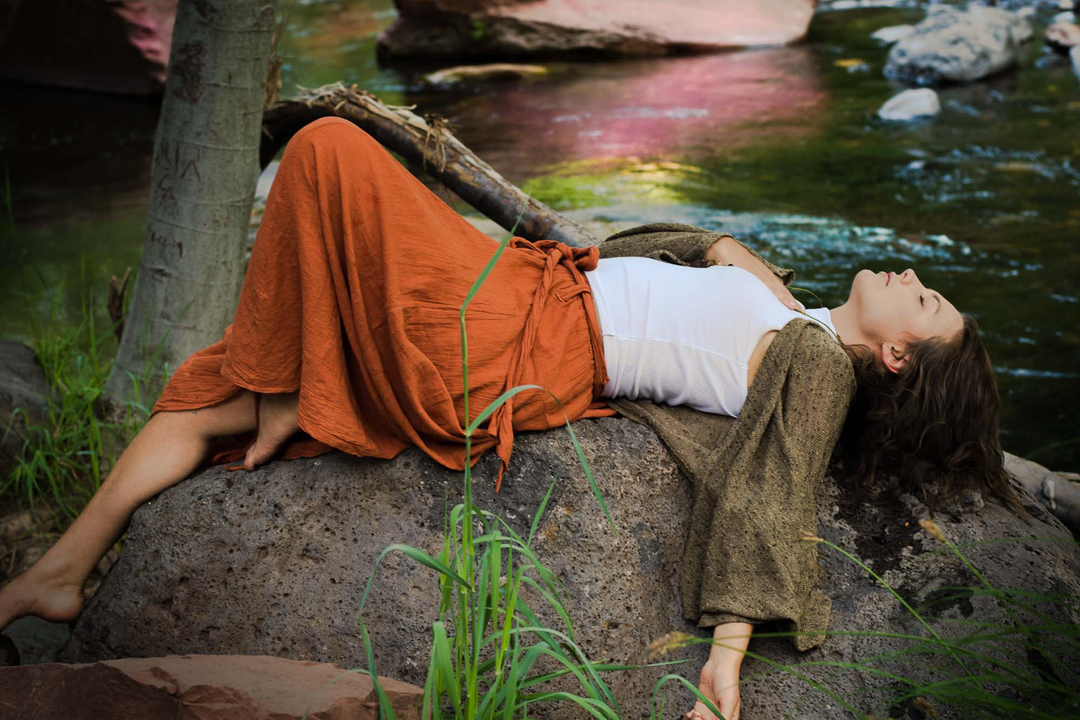 Female model is laying on a rock alongside a stream. She is wearing a white tank top, wrap and long orange skirt with sash. Her right hand is on her heart and eyes are closed.