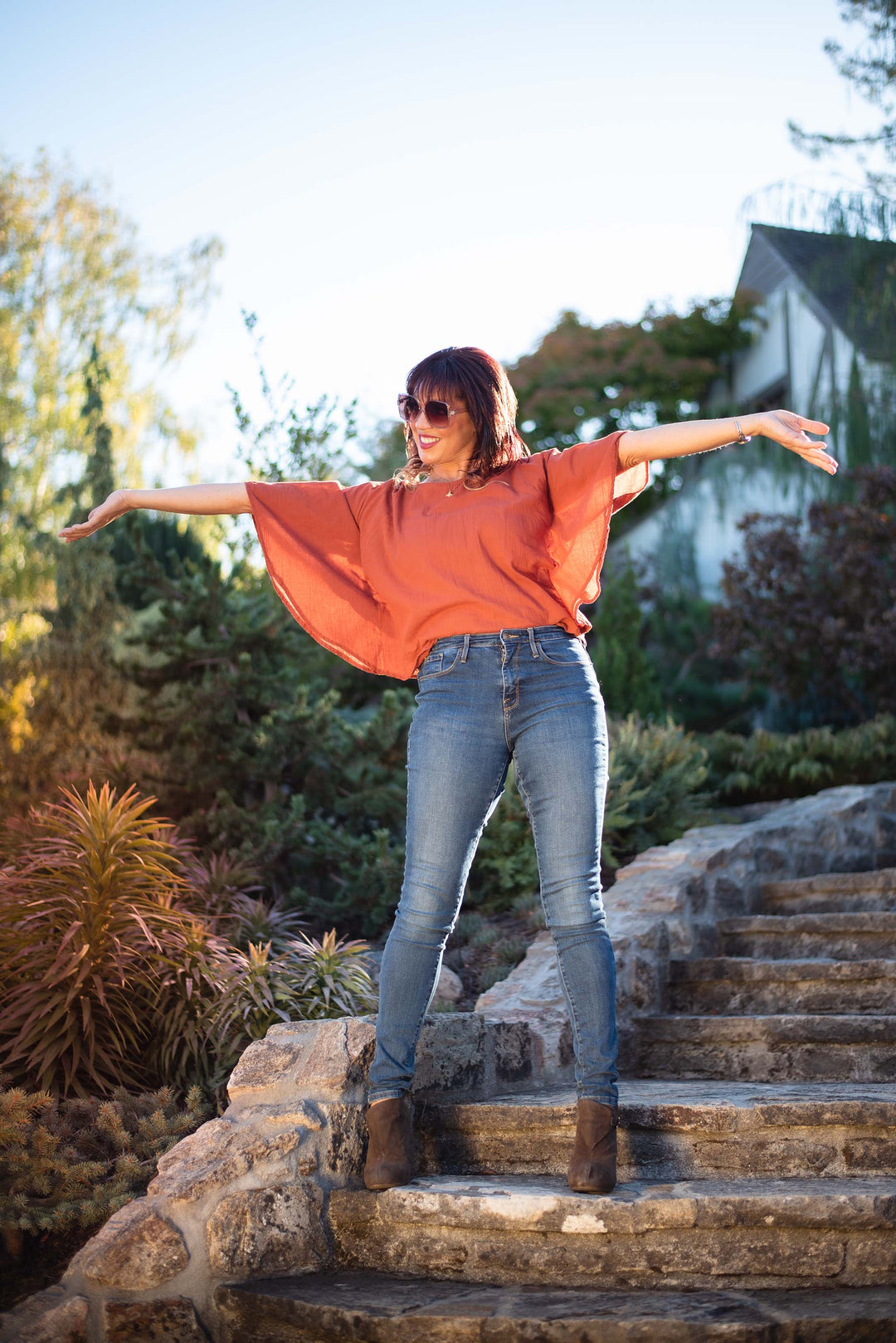 Model has on red winged top, blue jeans and brown booties. She is standing outside on stone staircase.