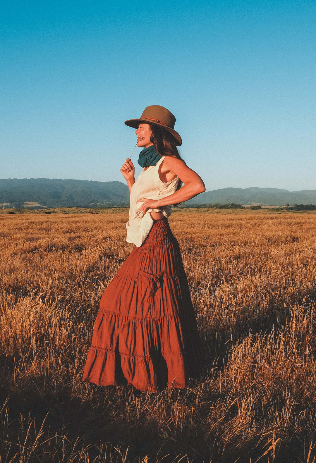 Woman adorned in long red skirt with pocket, tank top, scarf and cowboy hat stands in field at sunset.