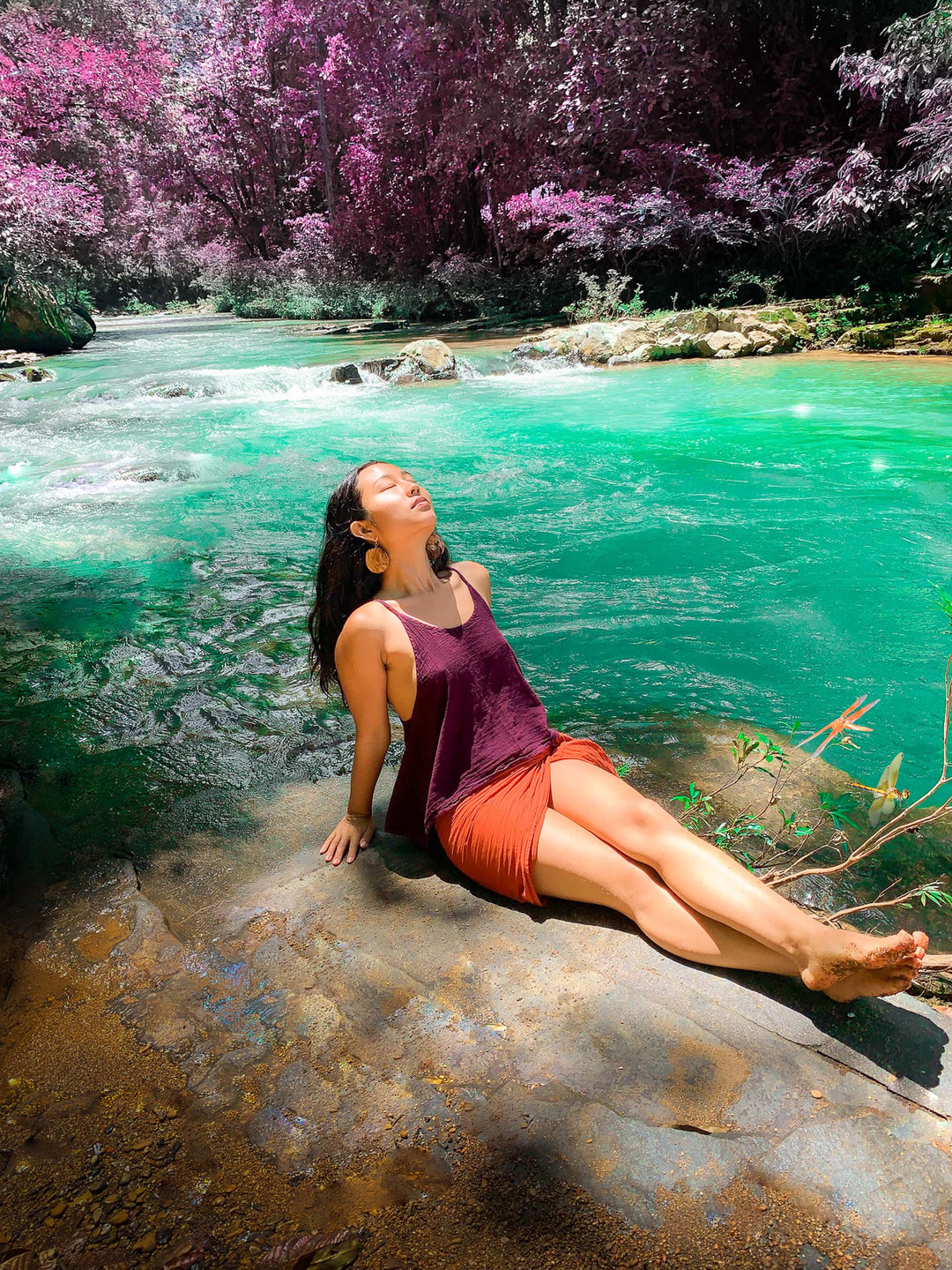 Woman has red wrap tied as a sarong skirt paired with purple tank top. She relaxes on a rock by turquoise water.