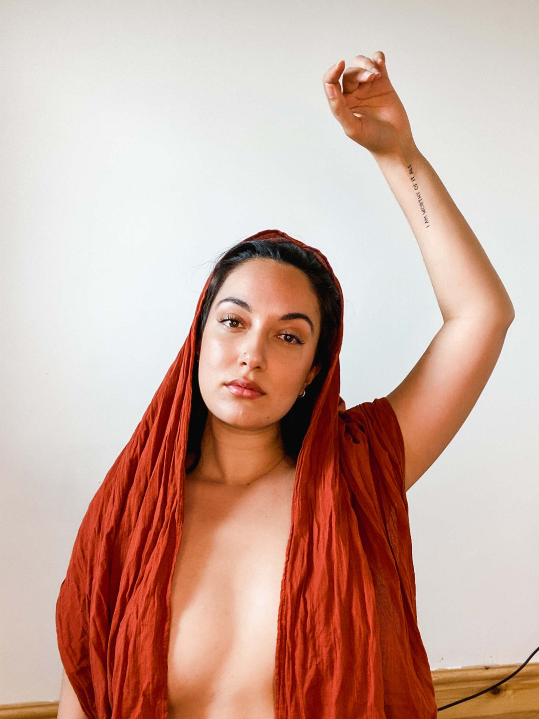 Woman is clothed in long red wrap that drapes over her head, shoulders and chest. Her left arm is in the air above her head.