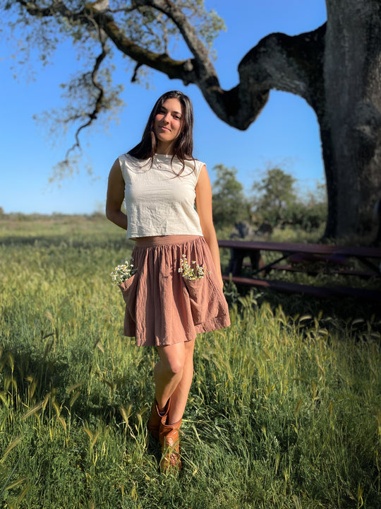 Female model wears un-dyed crop top and short pink skirt with pockets. White flowers peek out of the two pockets. She is standing under a tree in tall grass near a picnic table.