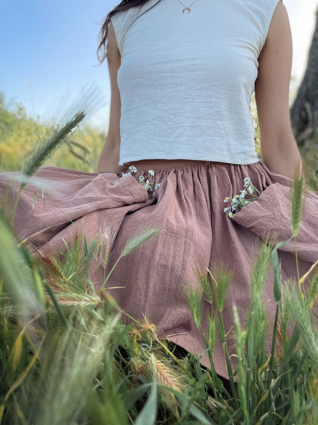 Model wearing natural white tank top and pink gauze short skirt is standing in tall grass. Skirt pockets have white flowers in them.