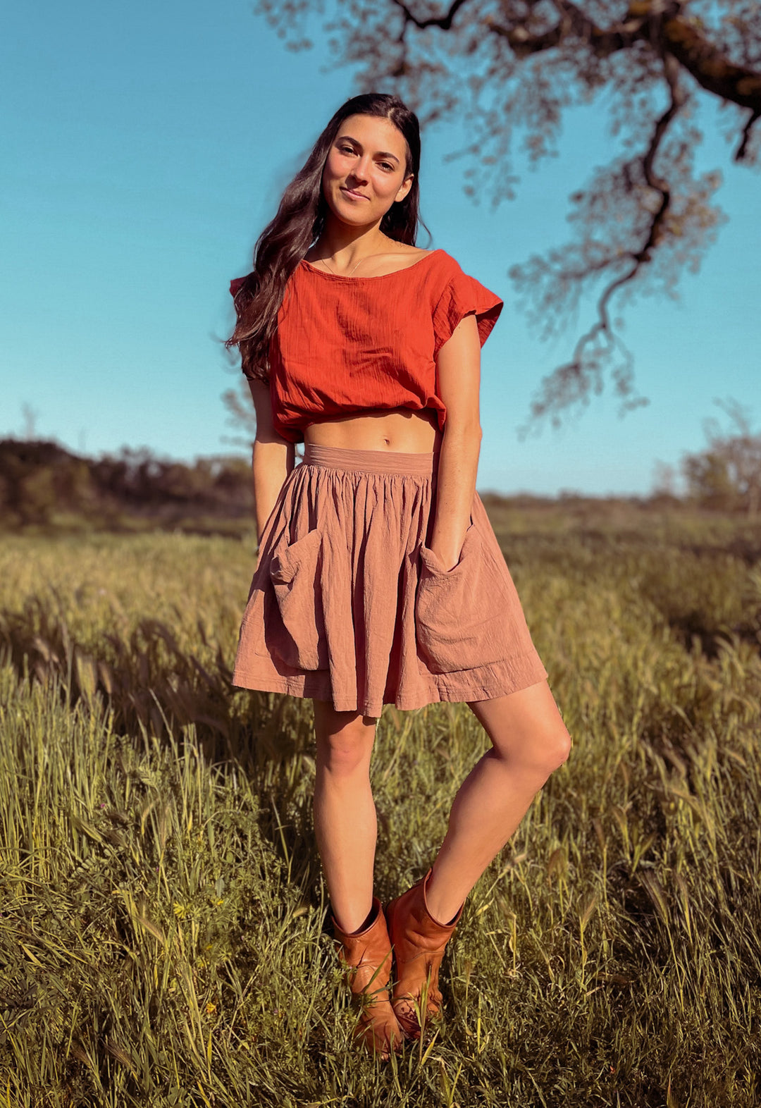 Woman stands outside in tall grass dressed in red blousy crop top paired with rose mini skirt and brown booties.