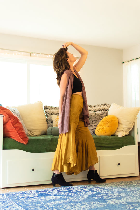 Woman standing in front of a day bed wearing flowing purple vest over a black crop top, gold-color bell bottom pants and black ankle boots. 