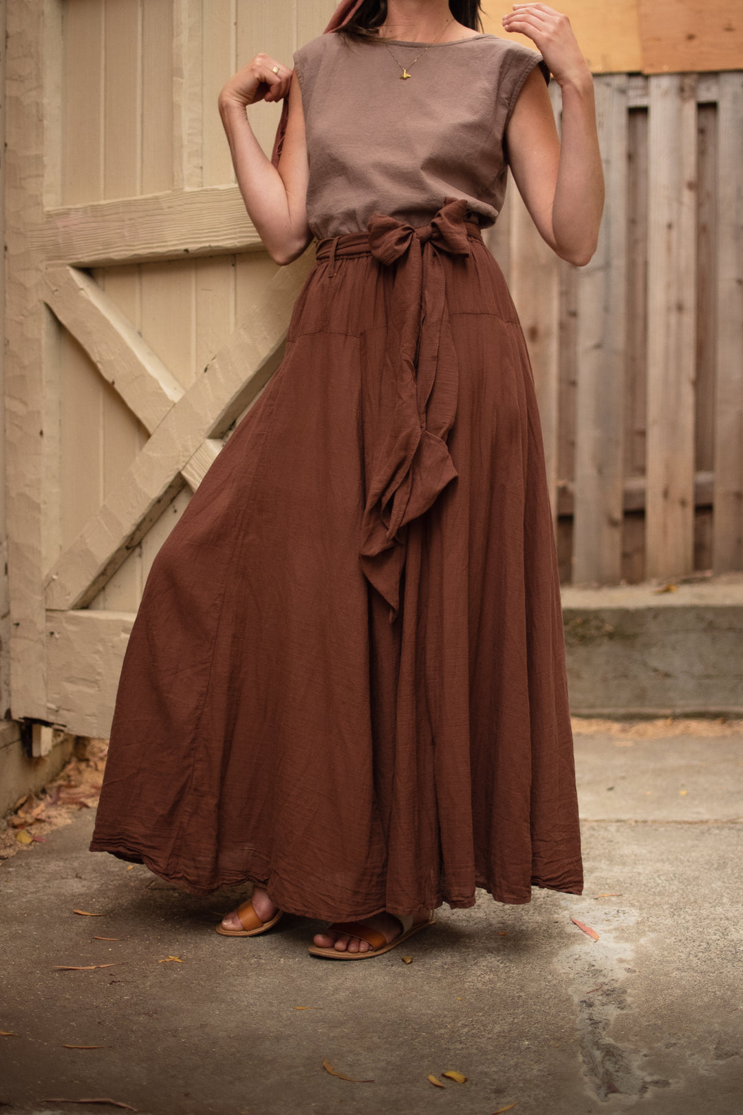 Female model is standing by barn door with light brown tank top and long dark brown skirt with bow. She has sandals on.