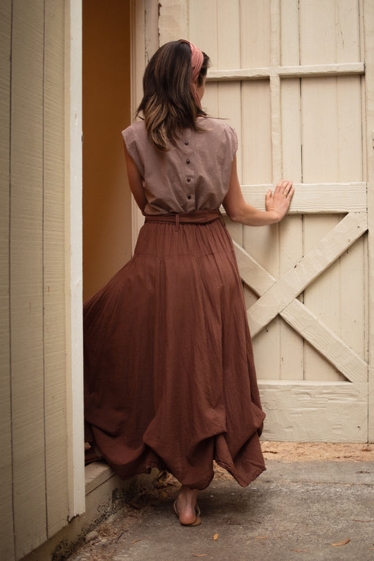 Back of female model standing at barn door. She wearing a tank top with buttons down the back and a long tiered brown skirt with sash. She has on sandals. Her right hand is on the door.