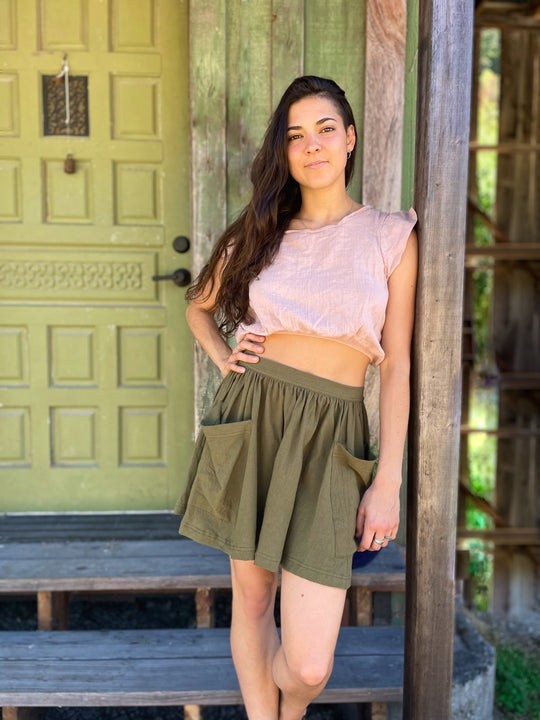 Woman leans against wooden post dressed in a light pink crop top with small sleeves and short green skirt with pockets.