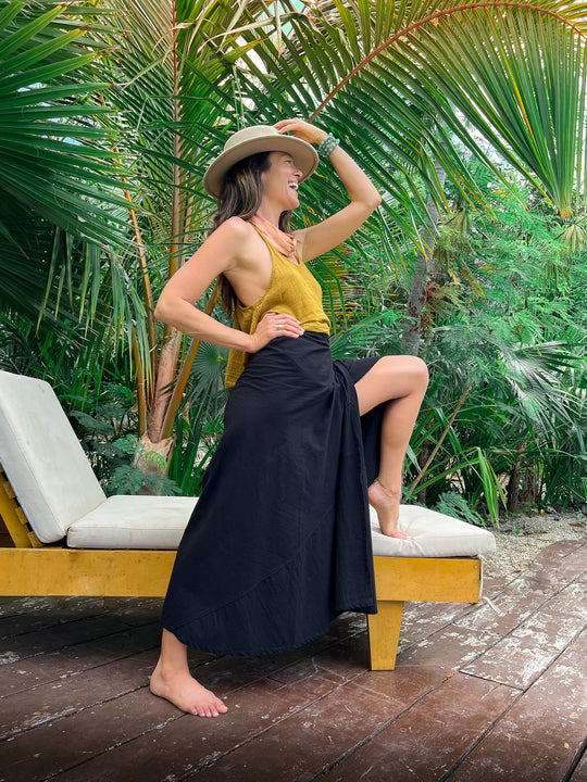 Model poses in jungle setting wearing black slitted skirt and gold tank top. She is barefoot.