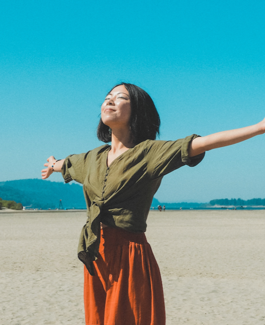 Woman wearing long-sleeve shirt with buttons and lounge pants holds her arms wide open.