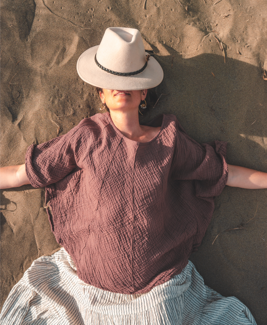 Woman relaxes on sand in full, blousy pink shirt with sun hat over eyes.