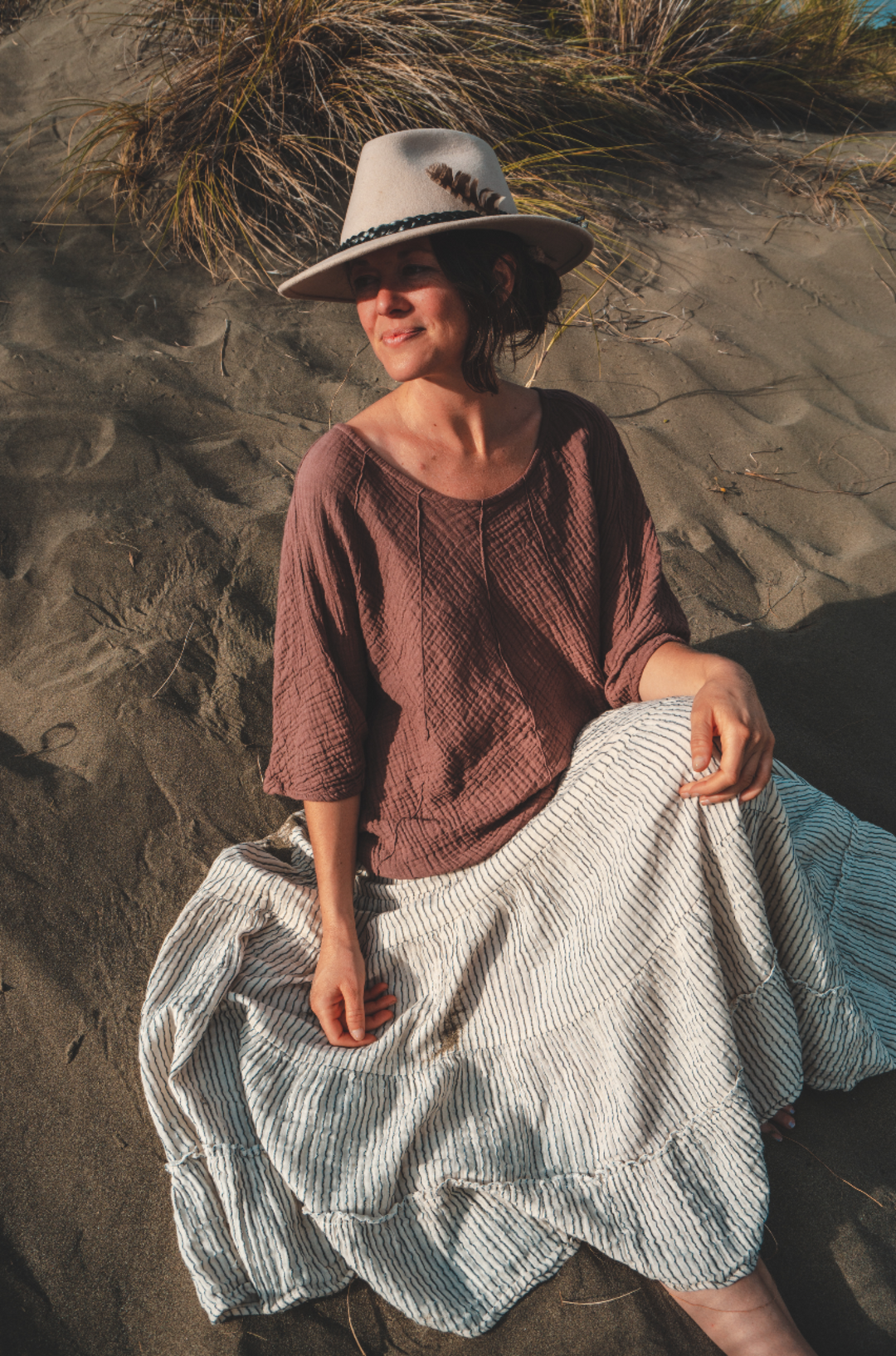 Model sits on sand dune in a tan hat, rose-color top and pinstriped skirt.