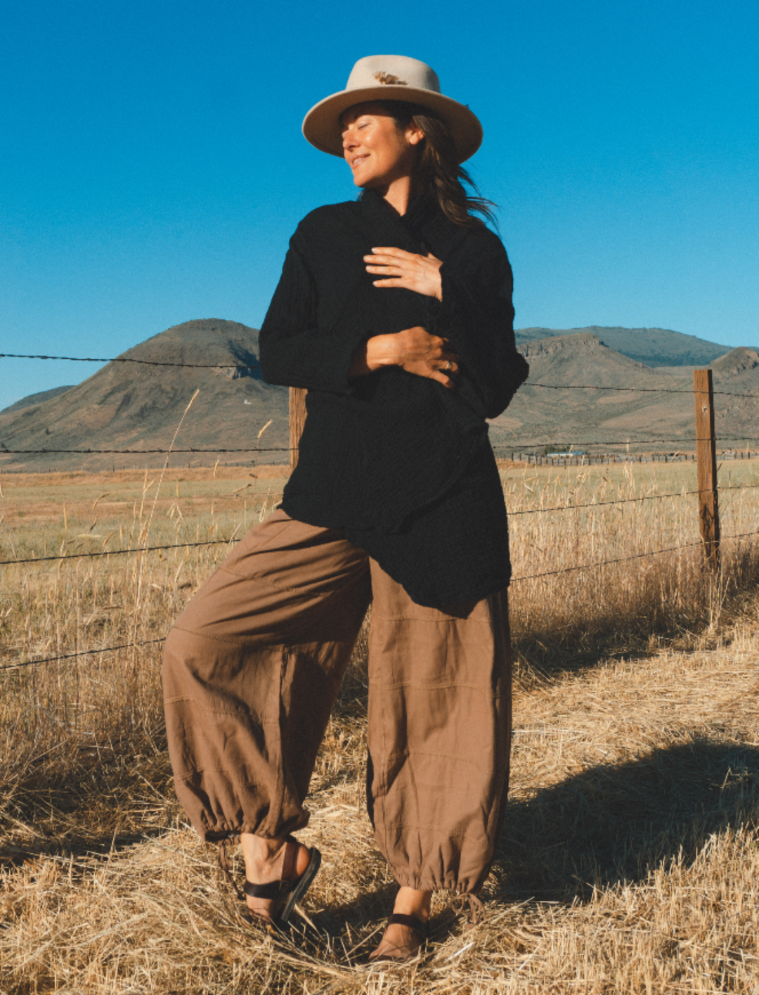 Woman stands in sun wearing tan felt hat, black cloak over white tank top and brown pants.